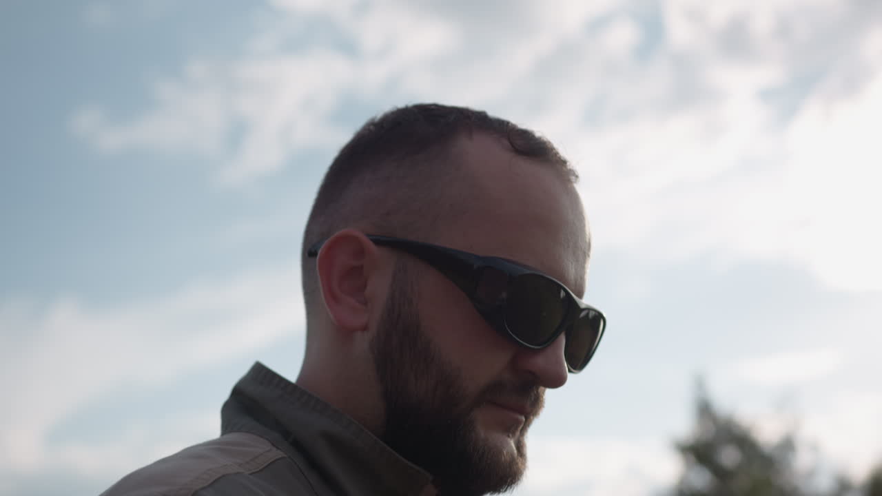 close up of man wearing dark sunglasses gazing into sky above blurred treetops, beard silhouette outlined against clouds, sun glare reflecting on lens, contemplative expression in outdoor nature