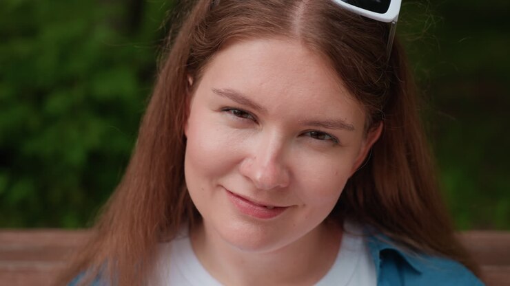 Close up of cute girl with white sunglasses on head turning slightly and smiling with elegance while relaxing on park bench, under daylight surrounded by lush green natural background