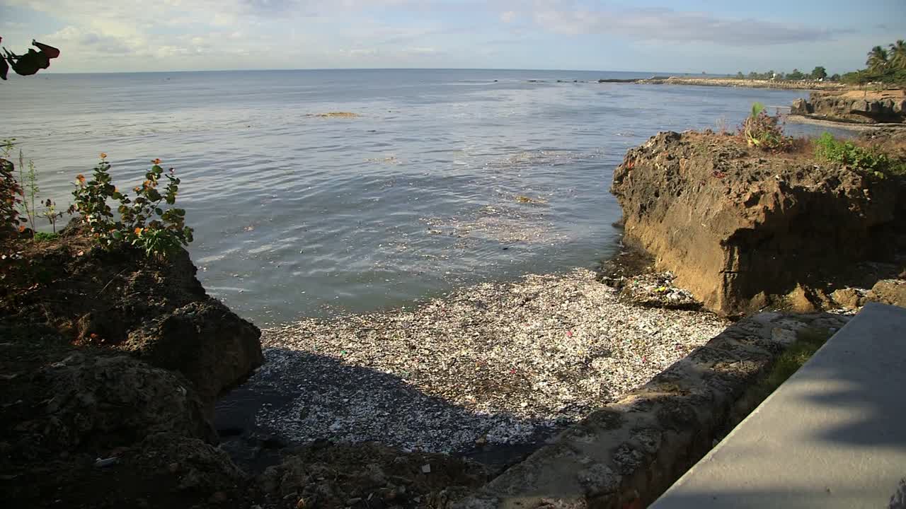 Klippefyldt caribisk kystlinje i Santo Domingo, Den Dominikanske Republik, med synlig plastikforurening, eroderet kystkant og roligt hav ved siden af en bynær havnefront