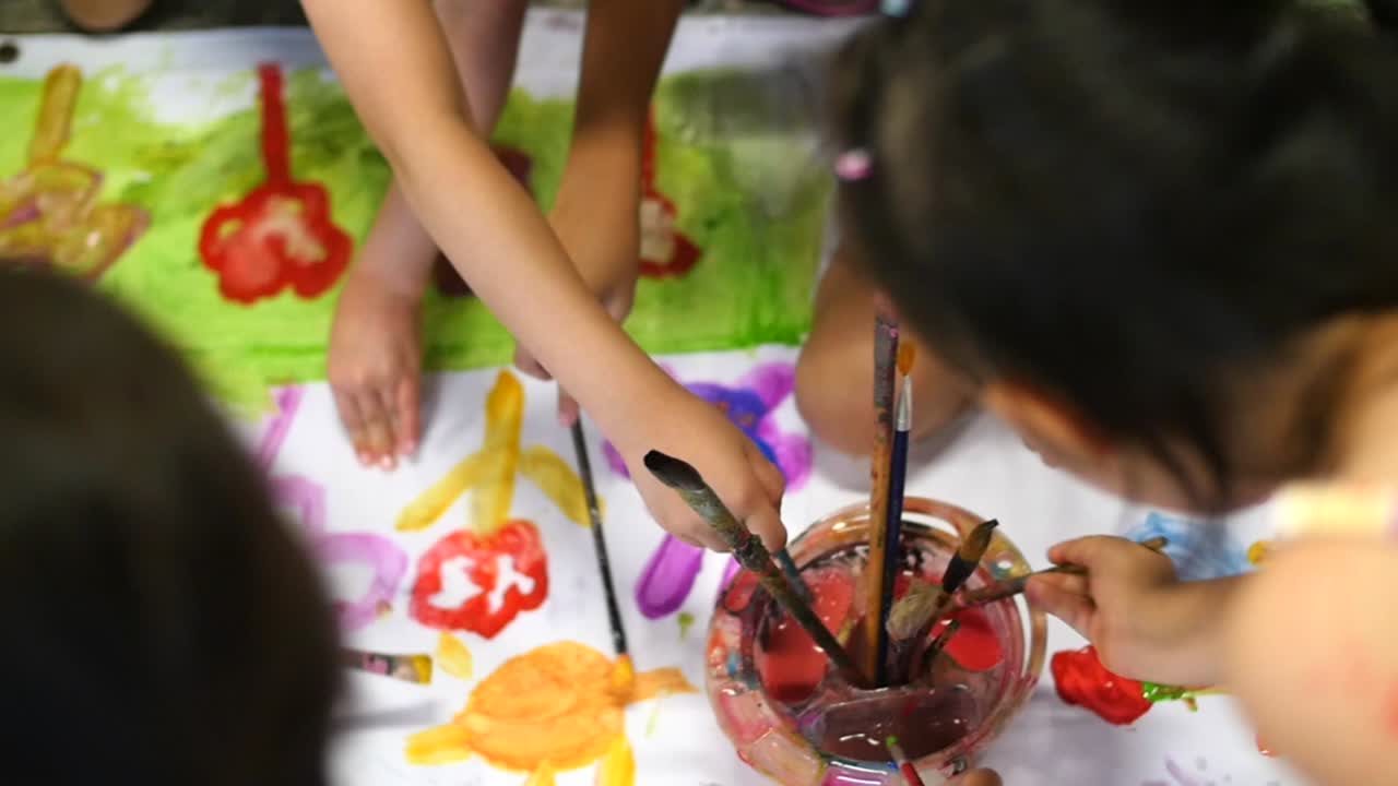 fotografía cenital de niños limpiando pinceles con agua y jadeando en una gran hoja blanca de papel filmada a cámara lenta