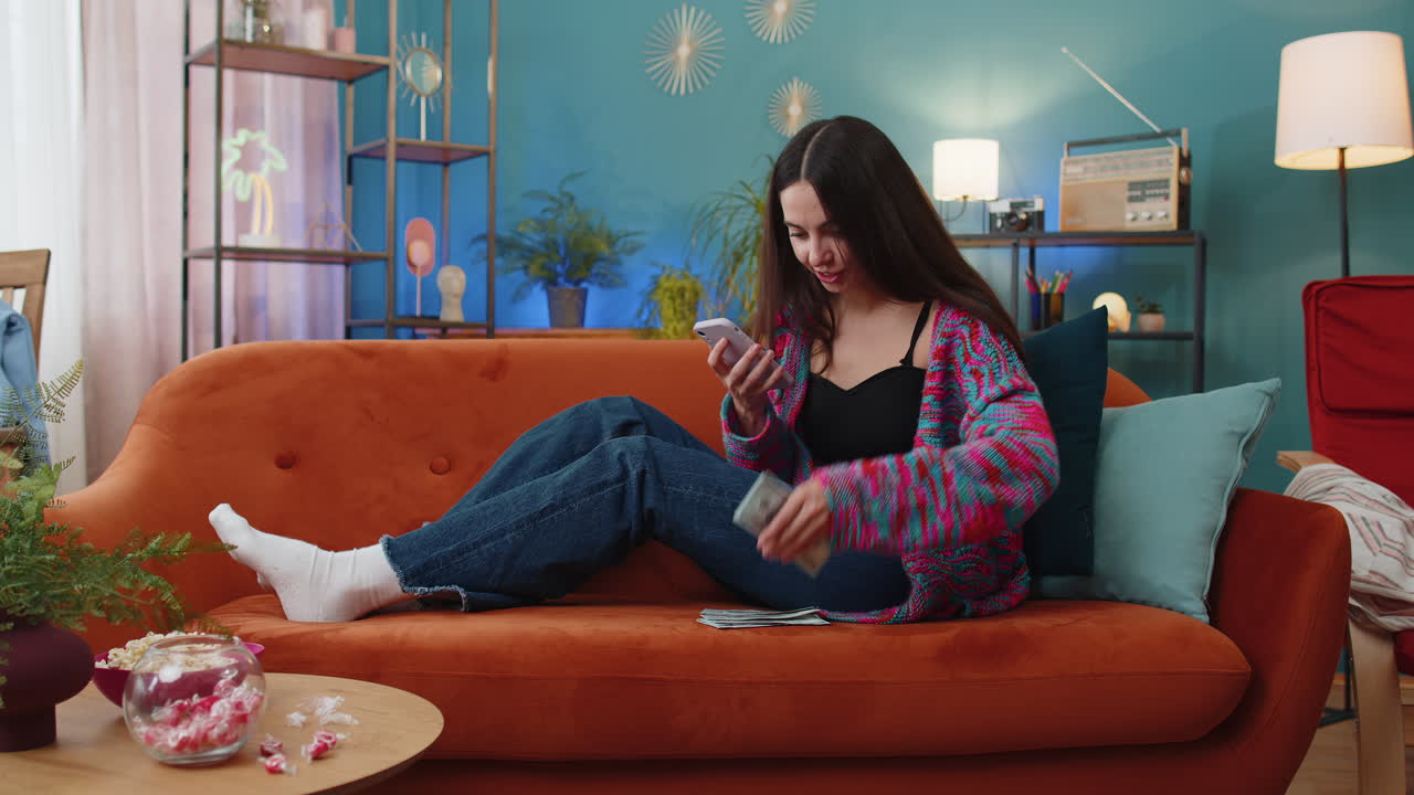 Smiling happy young woman girl counting money cash use smartphone income saves lottery win at home