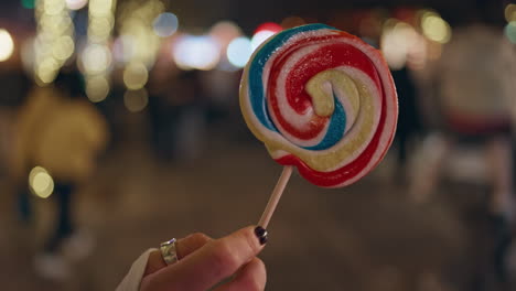 colorful lollipop woman hand at night amusement park closeup. unknown girl