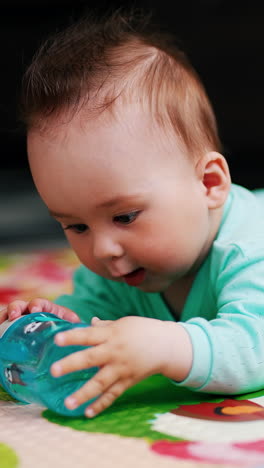 charming baby with funny hairstyle lies on mat. nice kid is playing with a bottle trying to take it in his mouth. close up. vertical video