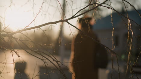 a back shot of a woman wearing a brown coat, jeans, and white shoes, carrying a backpack as she walks through a park. the foreground is dominated by sharp-focused tree branches