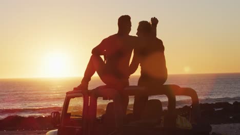 happy caucasian gay male couple sitting on car roof embracing at sunset on the beach