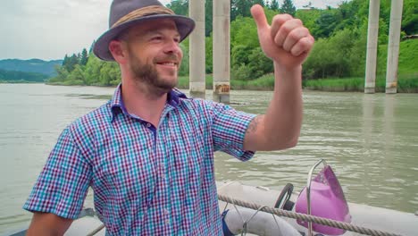 smiling man in hat gives thumbs up signal while on boat in river