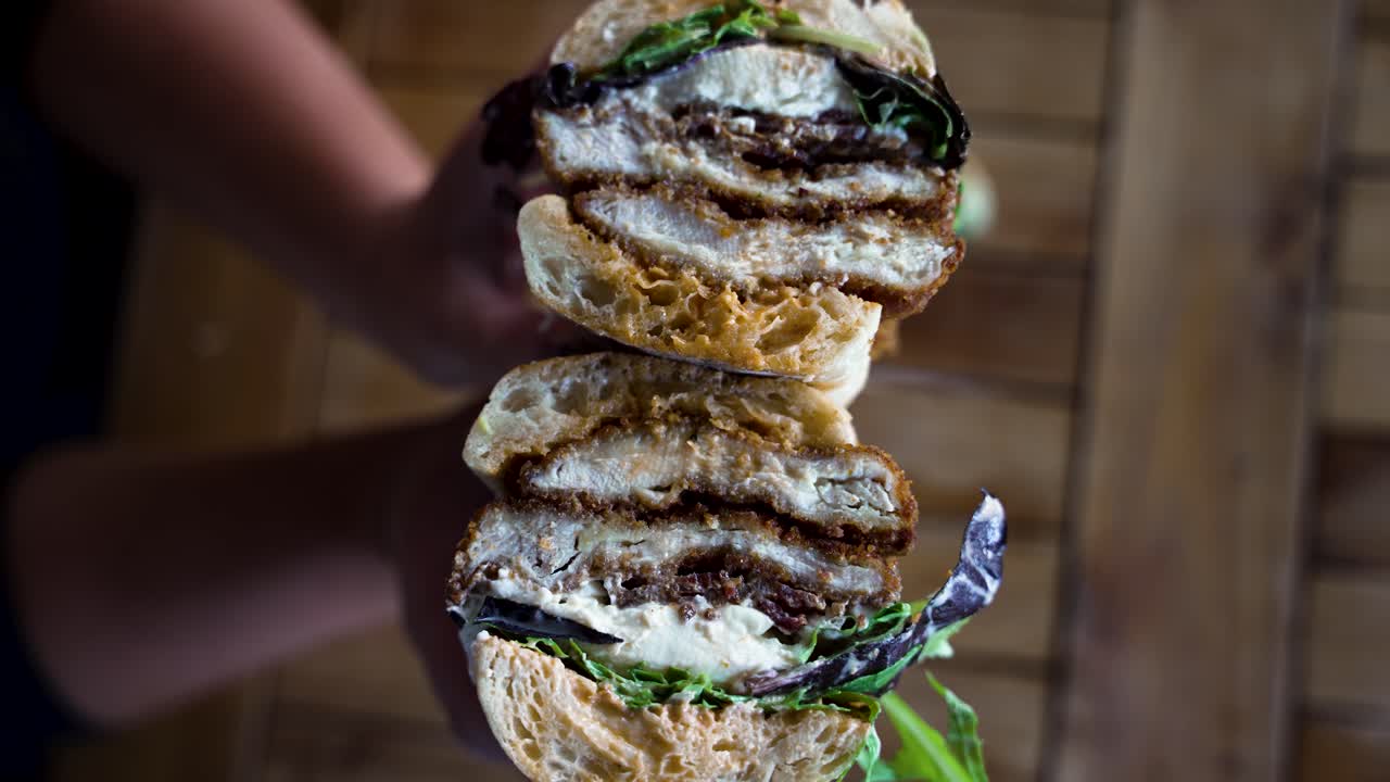 Close up in 4K of a chicken cutlet sandwich with lettuce and burrata on french brea, that's just been cut in half and opened to reveal the filling by hands with a perfect manicure