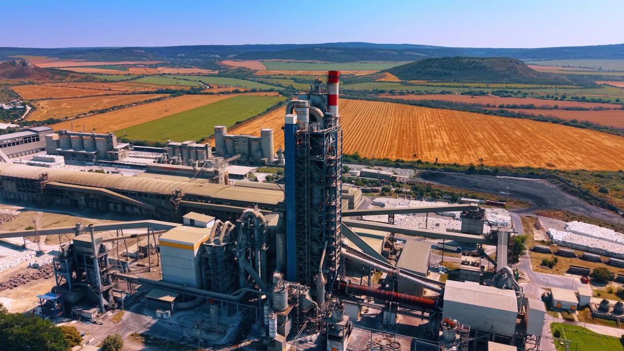 Cement production tower surrounded by harvested fields. Aerial perspective of a tall kiln structure, conveyors, silos and factory buildings within patchwork farmland.