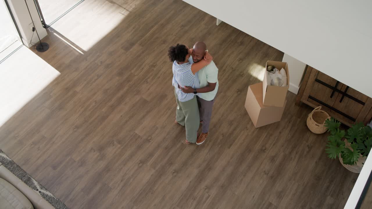 Diverse couple embracing as woman reaching, man lifting and swaying at home near move-in boxes