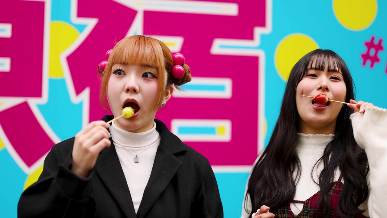 To japanske kvinder nyder farverige kandiserede frugtspyd mod en levende grafisk væg i Harajuku