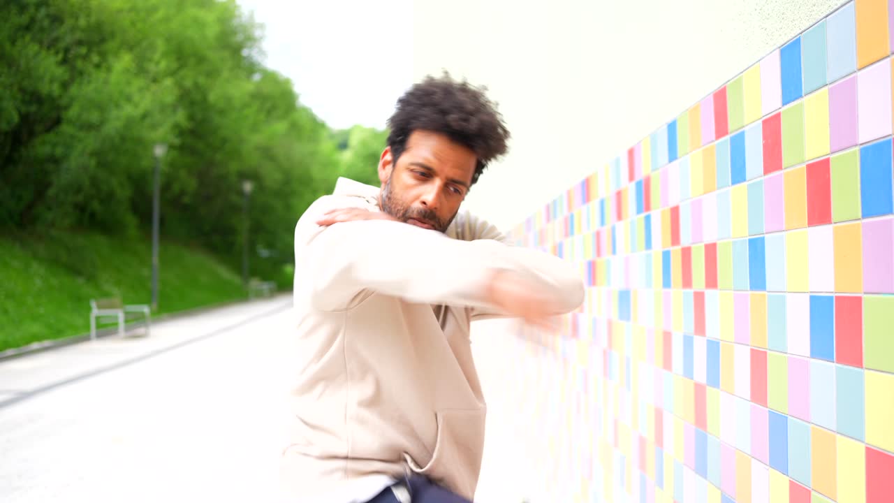 A man in a beige hoodie interacts with a vibrant, colorful tiled wall, posing, dancing, and expressing himself in an outdoor urban setting