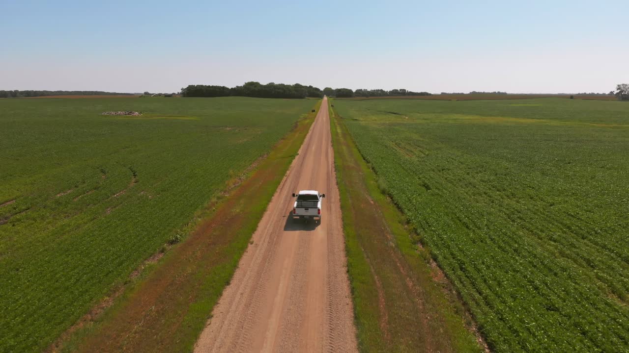 Aerial shot tracking a white pickup truck driving straight down a long dirt road between wide green farm fields