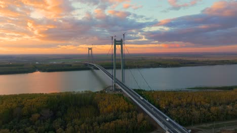 aerial close up shot showcasing a suspended bridge spanning a vast river, cars driving, after sunset orange colored sky, 4k50fps