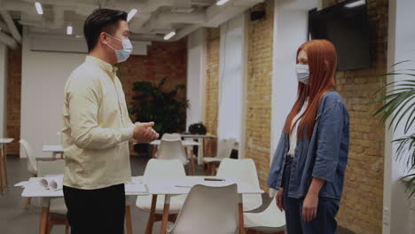 young woman and man with face masks do elbow bump, practice social distancing and talk about work with in the office