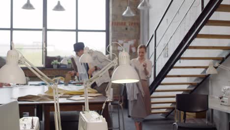 female fashion designer hanging dress on clothing rack in sewing workshop while her colleagues working in the background