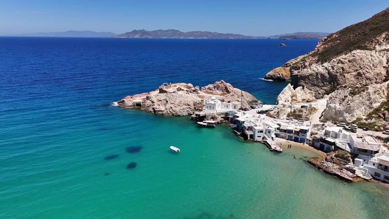Il villaggio di Firopotamos si snoda attorno a una baia turchese riparata mentre la telecamera aerea si allontana, rivelando case bianche, barche ormeggiate e la costa rocciosa di Milos.