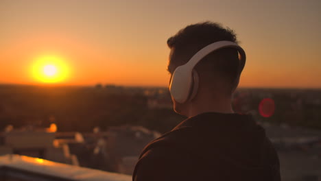 Un-Hombre-Camina-Sobre-El-Techo-Al-Atardecer-Con-Auriculares-Mirando-La-Ciudad-Desde-Lo-Alto-De-Un-Rascacielos-Al-Atardecer.-Relájate-Mientras-Escuchas-Música.-Disfruta-De-Una-Hermosa-Vista-De-La-Ciudad-Al-Atardecer-Desde-La-Azotea-Con-Auriculares