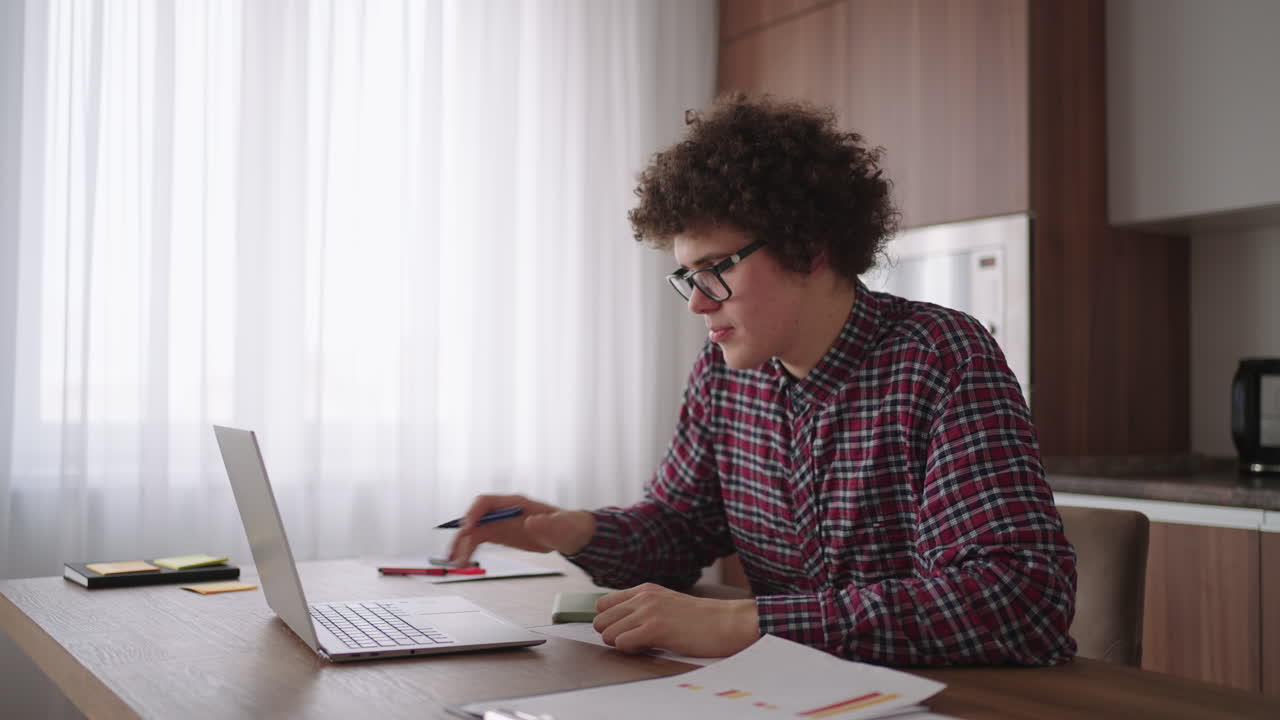 Curly haired Male student attractive young boy in glasses is studying at home using laptop typing writing in notebook. College student using laptop computer watching distance online learning seminar