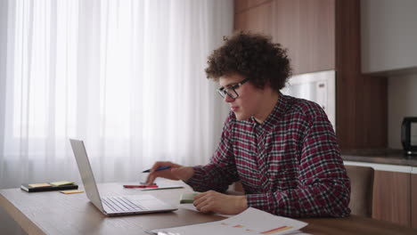 curly haired male student attractive young boy in glasses is studying at home using laptop typing writing in notebook. college student using laptop computer watching distance online learning seminar