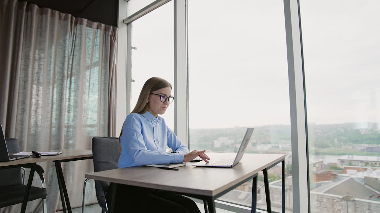Blonde long-haired woman in glasses working at computer sitting at desk. Light office background with panoramic windows.