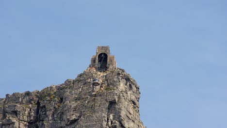 view from below of cable car ascending towards an arch structure atop towering cliff, set against clear blue sky in canada, dramatic scene highlights the height and ruggedness of mountain