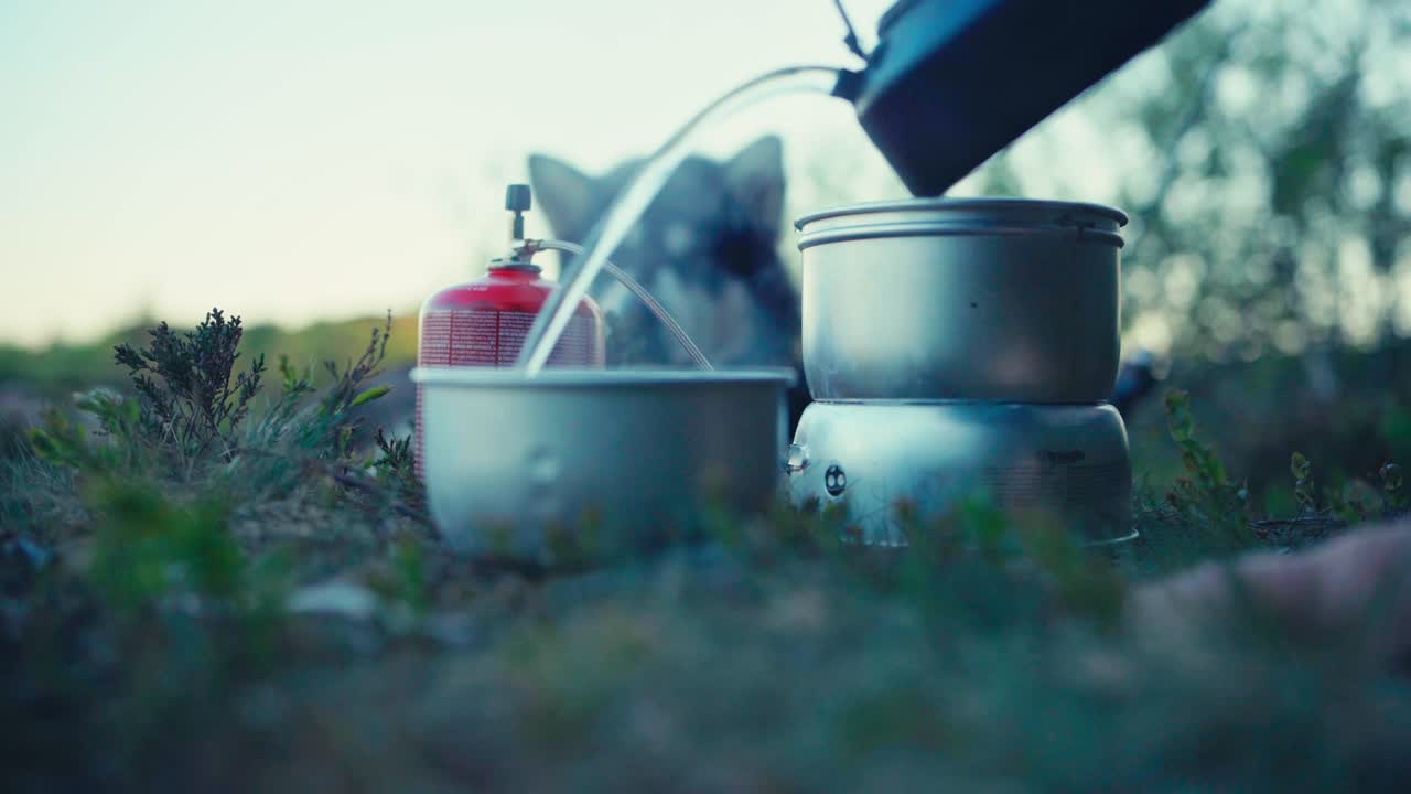 Pouring Hot Water from Kettle Into Pot On Grass At Campground