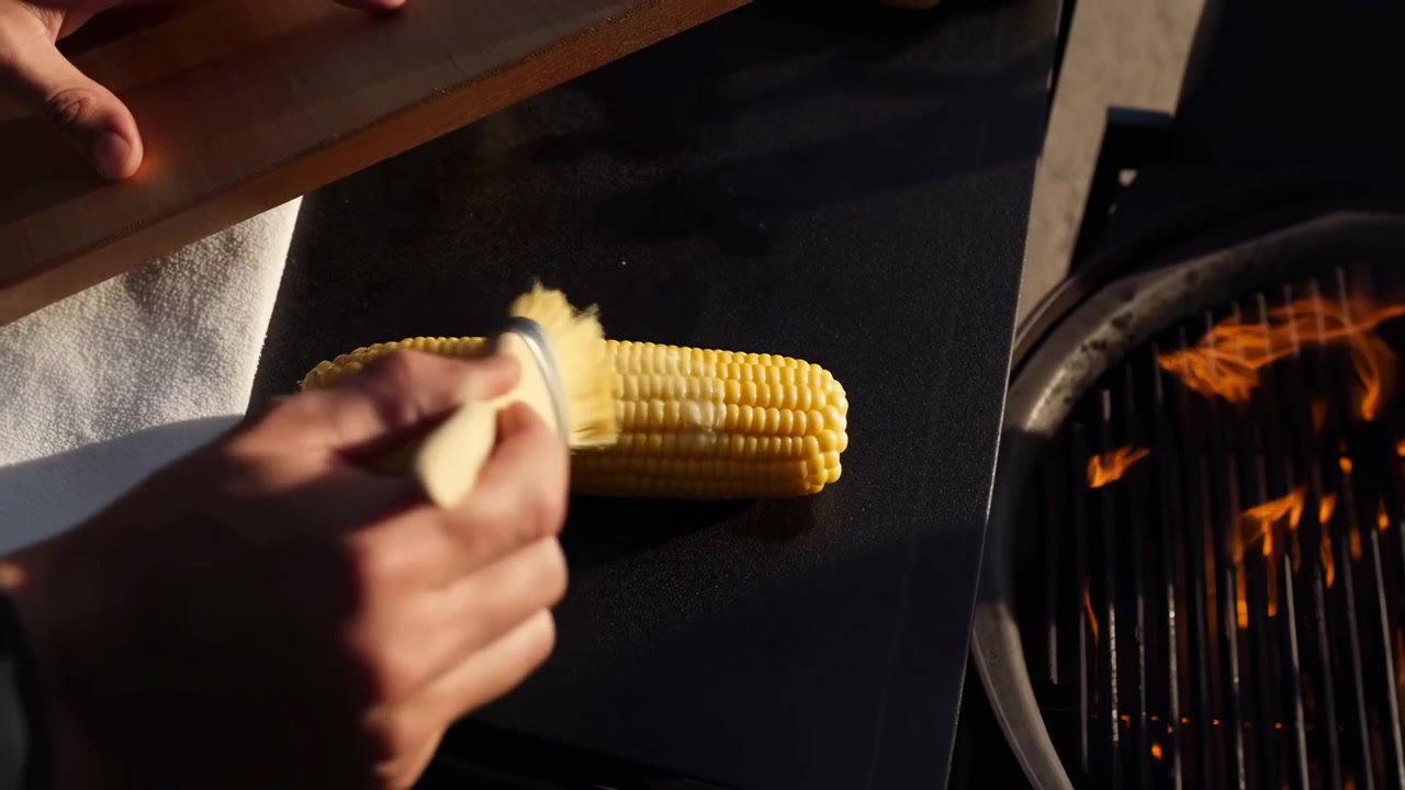Close-up of hands brushing butter or sauce over a corn cob on a dark barbecue surface, with bright flames glowing on the right and warm outdoor light creating