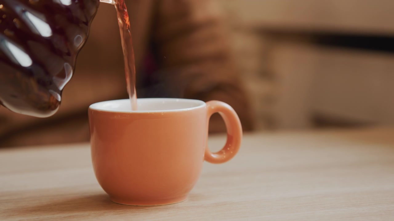 Primer plano de una mano vertiendo líquido oscuro en una taza de cerámica vacía de color melocotón sobre una mesa de madera. La acción es constante y enfocada, con el fondo suavemente difuminado para enfatizar el sujeto. La iluminación suave resalta la escena.