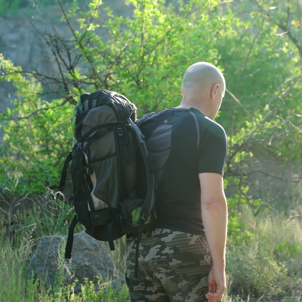 Back view, in movement, a bald hiker man with a backpack on his shoulders walking on a path near the green trees. Male backpacker hiking in mountain adventure in summer. Close-up