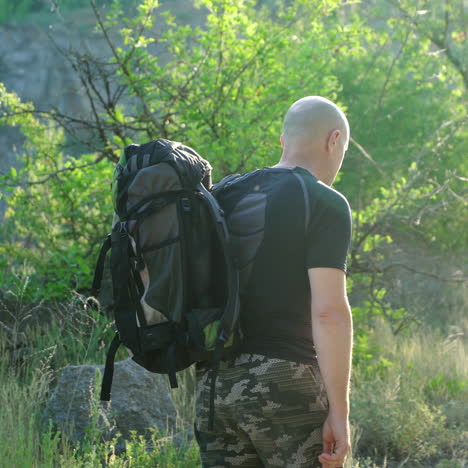 back view, in movement, a bald hiker man with a backpack on his shoulders walking on a path near the green trees. male backpacker hiking in mountain adventure in summer. close-up