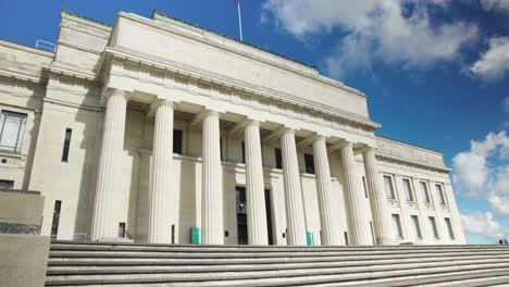 low angle pan right view of the auckland war memorial museum, its fluted columns, and the staircase leading up to it on a bright sunny day with clouds drifting through the blue sky