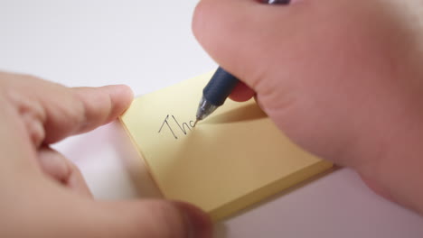 righthand writing the words "thank you" on a post-it paper pad while the lefthand holds it in place