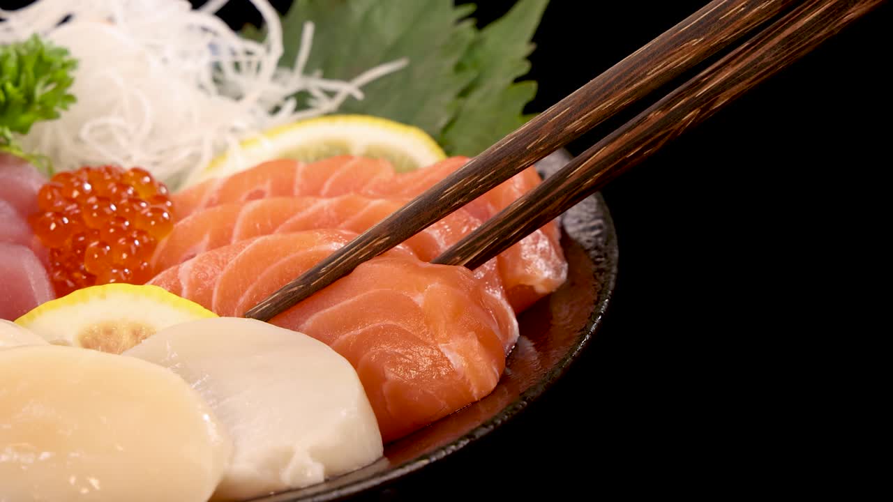Essstäbchen heben behutsam eine Scheibe Lachs-Sashimi von einem japanischen Sushi-Set, arrangiert auf schwarzem Hintergrund mit heller, gleichmäßiger Beleuchtung und Nahaufnahme-Bildausschnitt.