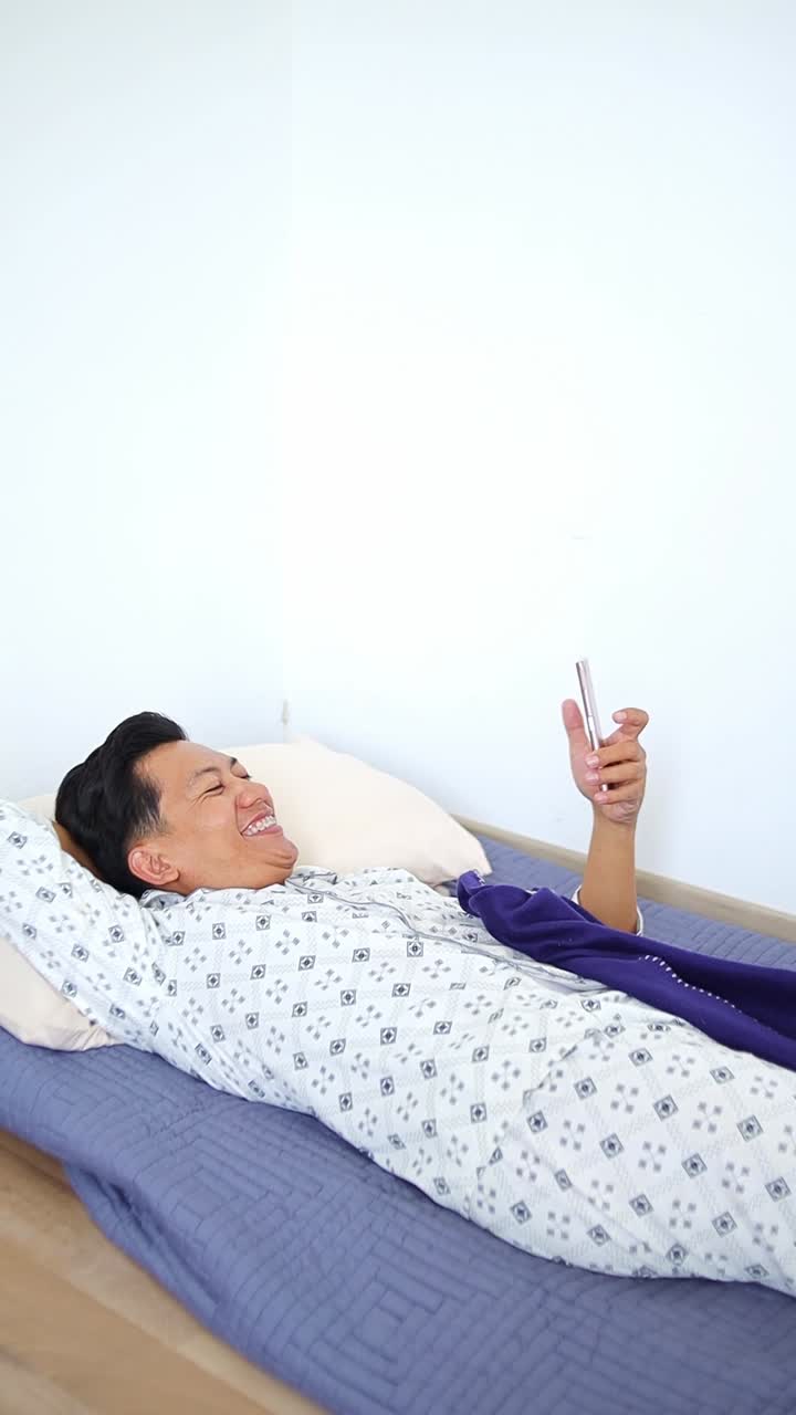Happy Young asian man in pyjamas laughs while scrolling social media on smart phone at bed room