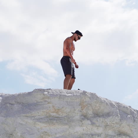 shirtless athlete in cap and glasses on mountain. view from below on a sportsman standing on the top of white hill.