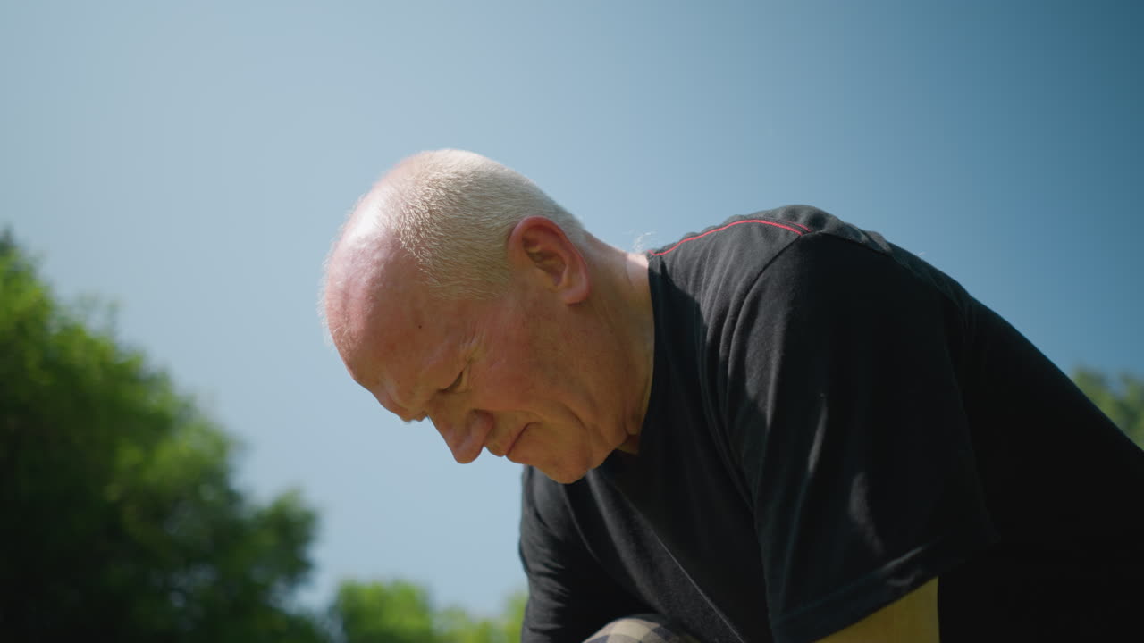 An elderly man kneels down, deeply focused on a task, and lifted his head slightly, the background features a blur of trees under a clear sky
