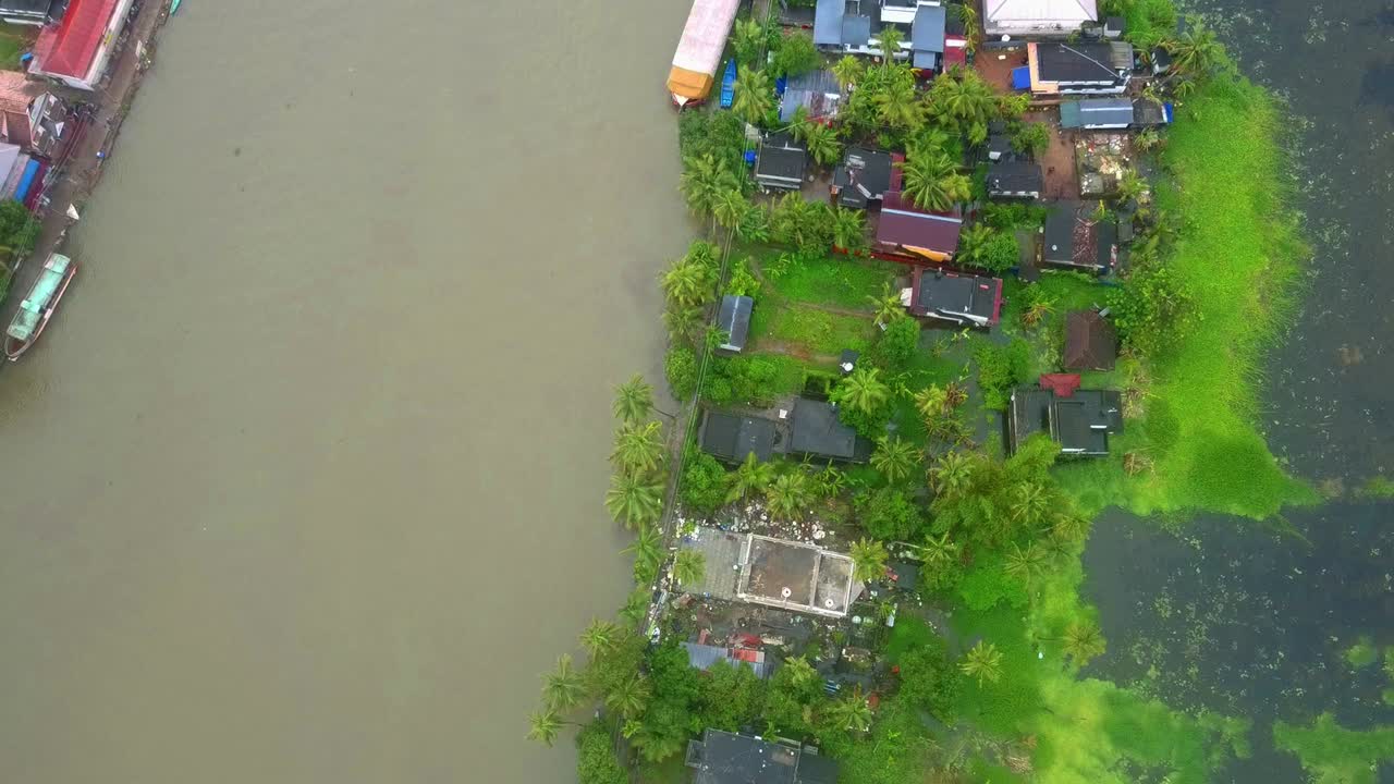 Aerial top-down view of a tropical village settlement bordering a muddy backwater canal, showing debris from a demolished damaged house and lush palm trees in Kerala, India