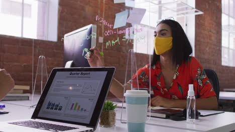 Mujer-De-Negocios-De-Raza-Mixta-Escribiendo-En-Una-Pantalla-De-Estornudo-Sentada-Frente-A-Una-Computadora-Con-Mascarilla