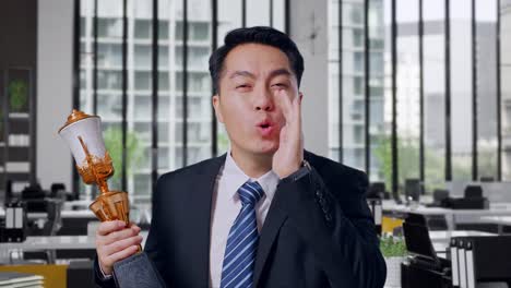 close up of asian business man in a suit and tie with a gold medal holding a gold trophy and yelling positively with hand over mouth in office