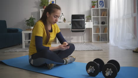 Una-Joven-Adicta-A-Internet-Está-Viendo-Redes-Sociales-En-Un-Teléfono-Móvil-Sentada-En-El-Suelo-De-La-Sala-De-Estar-Durante-El-Entrenamiento-Deportivo-Con-Equipo-Deportivo-En-Casa