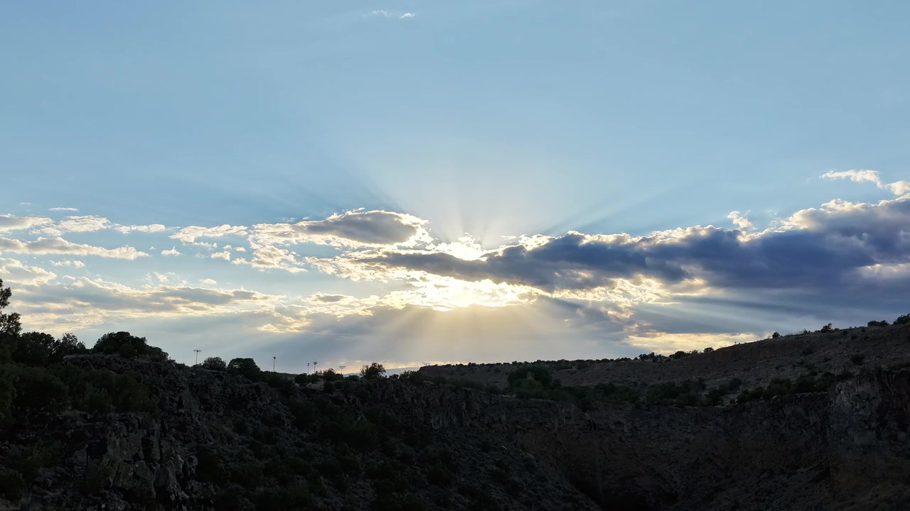 4K drone timelapse pushing forward towards a rugged canyon cliff edge. The shimmering evening sun breaks through scattered clouds, casting radiant sunbeams across the desert landscape.