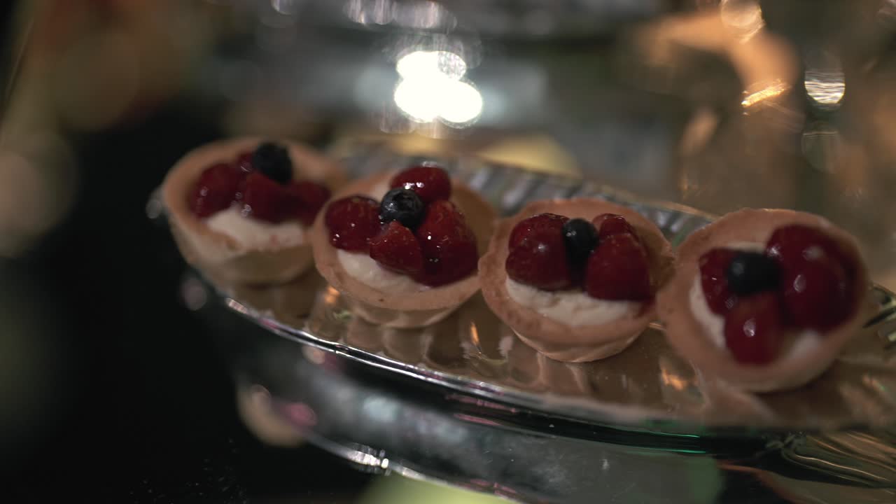 Sølvbakke på et spejlbeklædt bord med bagte dejkopper med fløde og glaserede bær. Festlys reflekteres på overfladen ved denne elegante bryllupsdessertbuffet