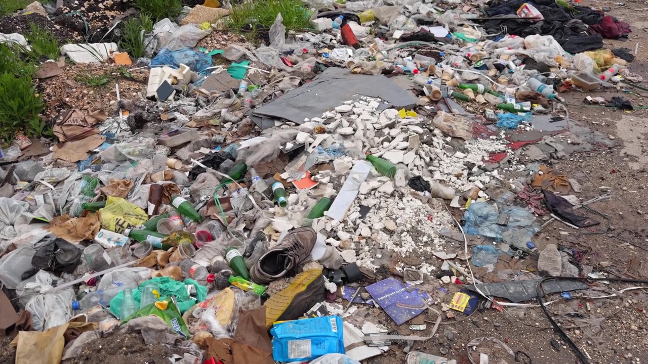 4K cinematic close-up shot of heavy plastic pollution, glass bottles, and construction debris piled on an illegal outdoor dump site. Highlights environmental and ecological waste issues.