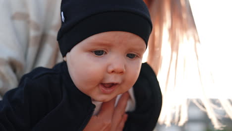 face of an adorable caucasian baby with grey eyes. kid in black cap in mom's hands outdoors. close up.