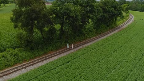 an aerial view of two amish girl walking on a single rail road track in the middle of amish farm lands