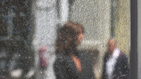 woman standing motionless behind cracked glass panel, experiencing urban anonymity while blurred pedestrians passing by on bustling metropolitan street