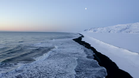 AERIAL:-Flying-over-Black-Beach-with-white-arctic-snow-mountains-in-background-in-Iceland-in-Winter-Snow,-Ice,-Waves,-Water