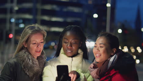 group of female friends on city street at night ordering taxi using mobile phone app