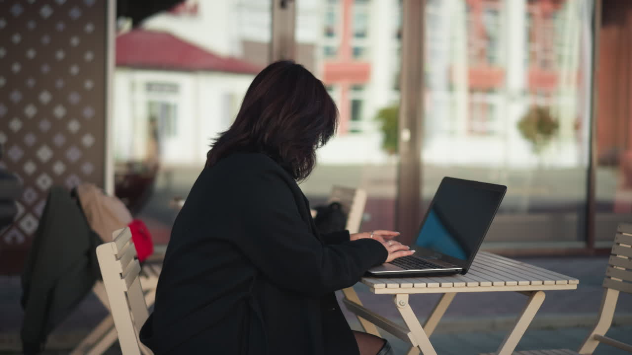 Professional woman working on laptop outdoors with coffee cup on table, decorative black pillar nearby, and blurred urban background of glass and buildings
