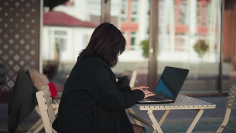 professional woman working on laptop outdoors with coffee cup on table, decorative black pillar nearby, and blurred urban background of glass and buildings